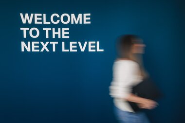 Mitarbeiterin geht an einer Wand mit dem Schriftzug "Welcome to the next level" vorbei | © Johannes Radlwimmer